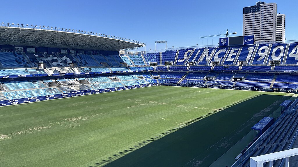 View of Estadio La Rosaleda in Málaga, Spain, featuring blue seating with "SINCE 1904" displayed, a partially covered stand, and a sunlit football pitch with visible wear.