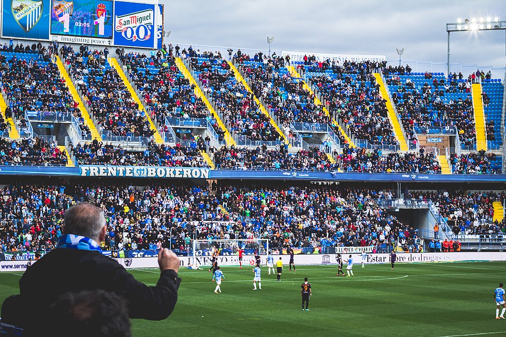 A lively scene at Estadio La Rosaleda in Málaga, Spain, showing fans in the stands, a digital scoreboard, and players on the pitch during a football match, with the "Frente Bokerón" supporters' banner visible.