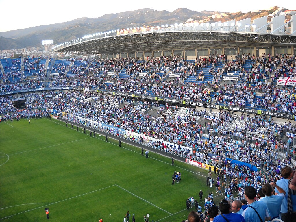 Estadio La Rosaleda in Málaga, Spain, with fans celebrating in the stands and on the pitch, under stadium lights, with mountains visible in the background.