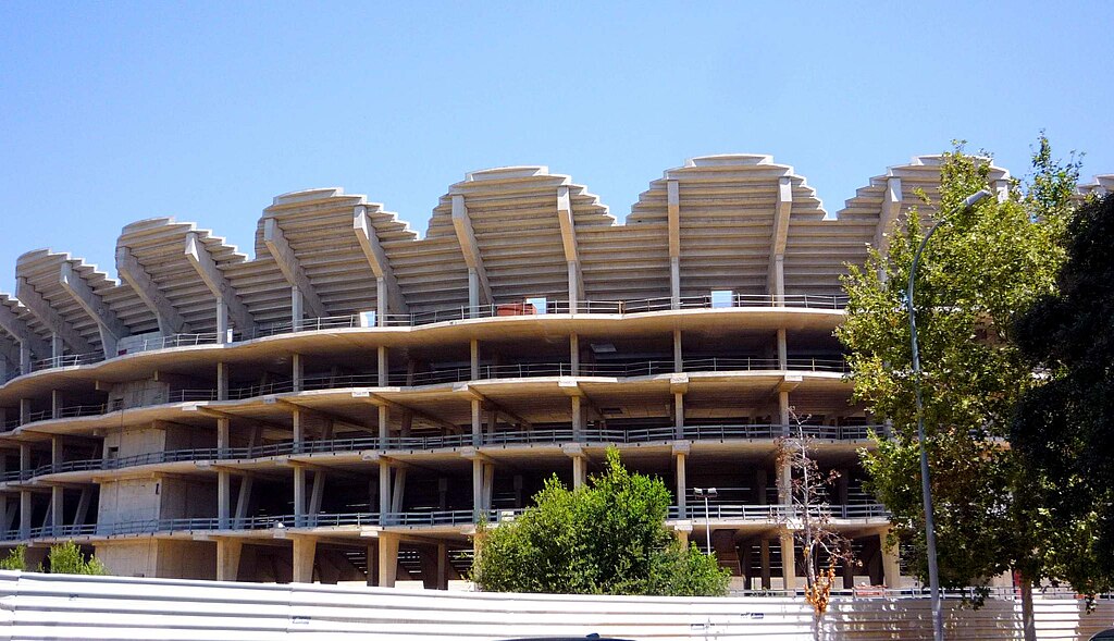 Partially constructed Nou Mestalla stadium in Valencia, Spain, featuring an unfinished concrete structure with distinctive layered roofing, surrounded by trees and a clear blue sky.