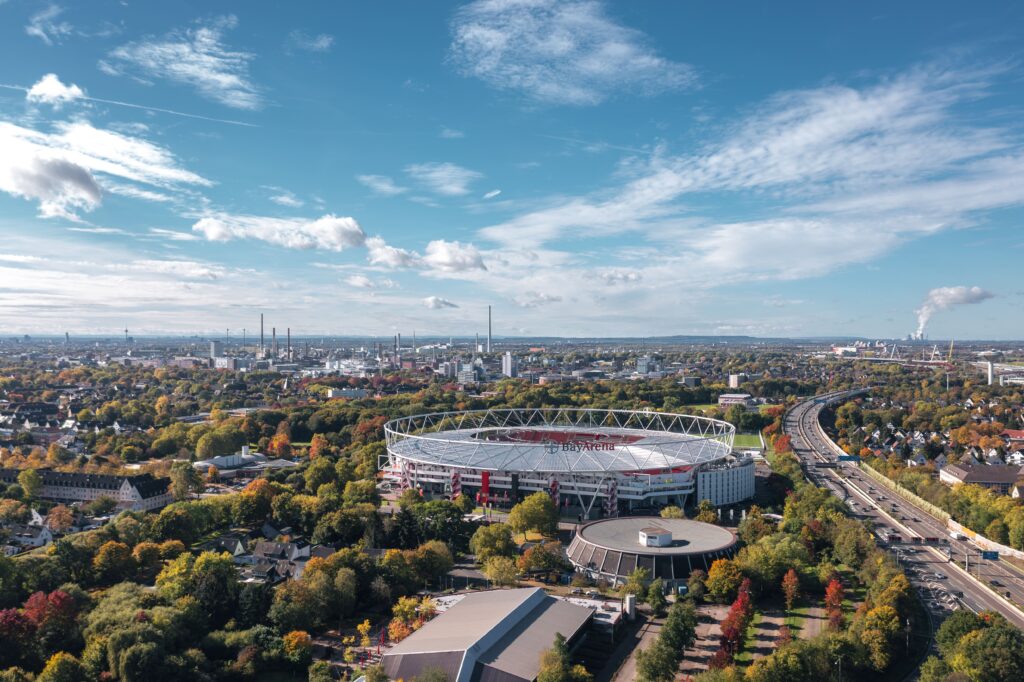 Aerial view of BayArena in Leverkusen, surrounded by greenery, roads, and the city skyline in the background.