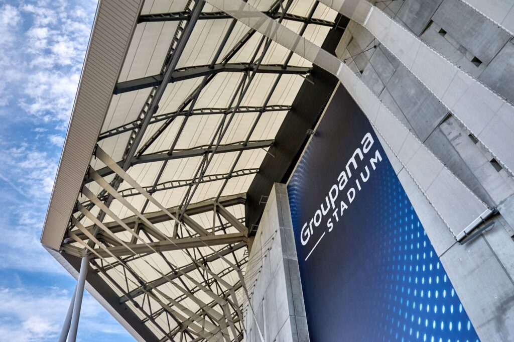 Uma vista arquitetônica moderna do Estádio Groupama, com sua estrutura de aço elegante, painéis de teto brancos e uma placa azul destacada do estádio.