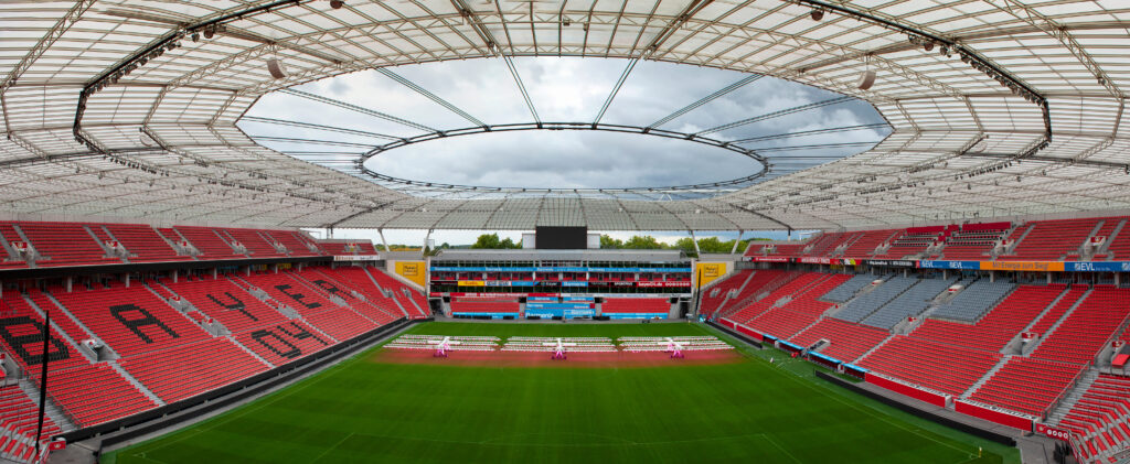 Interior view of BayArena, home of Bayer Leverkusen, with red seating, a green pitch, and a partially covered roof.