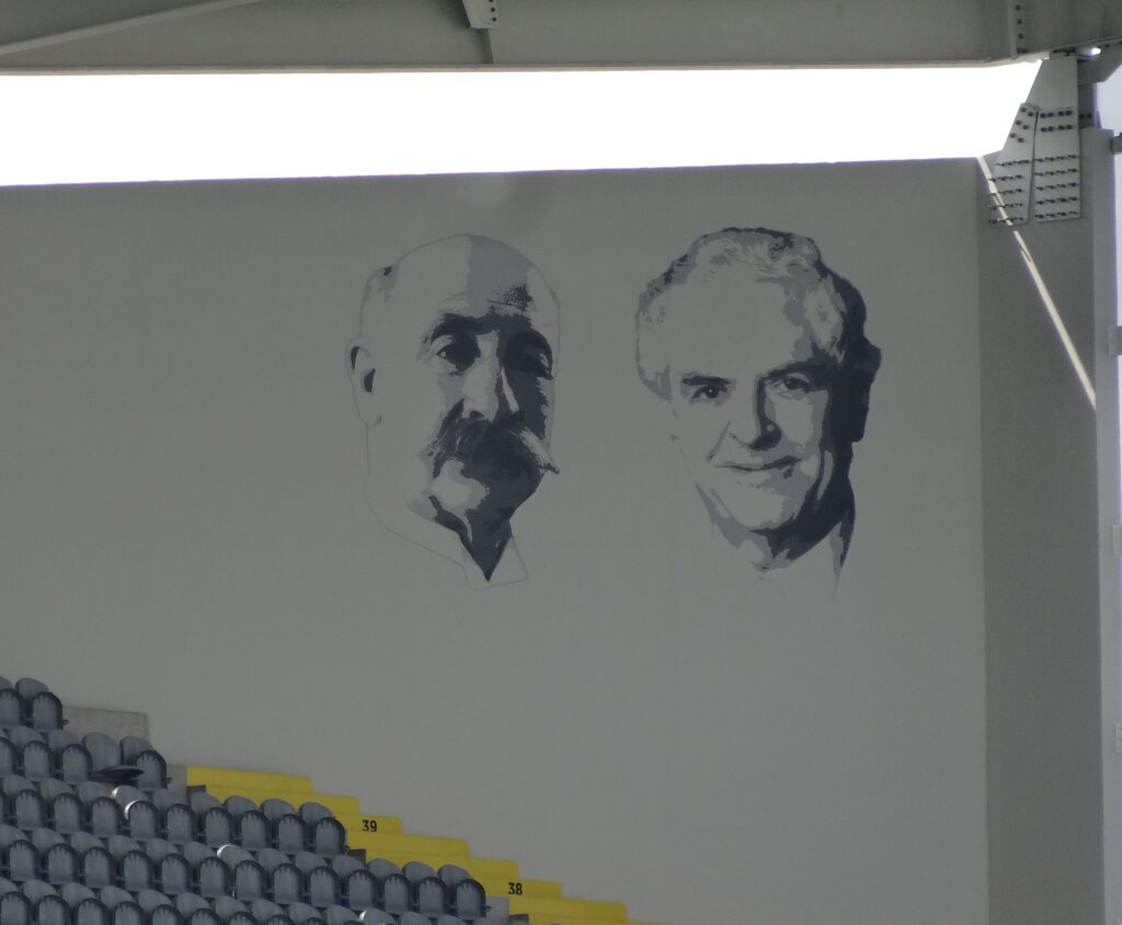 Wall mural inside Stade Félix Bollaert-Delelis featuring portraits of Félix Bollaert and André Delelis, commemorating their contributions to the history of the stadium and the region. The artwork is located above rows of empty seating in the stadium.