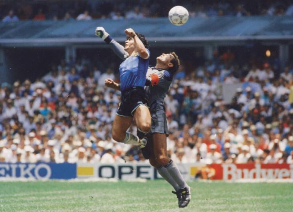 Football action moment with a player and a goalkeeper jumping to contest the ball in a packed stadium.