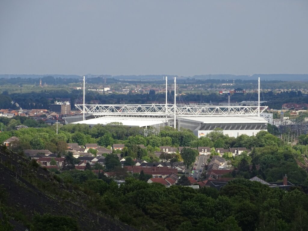 A distant view of Stade Félix Bollaert-Delelis in Lens, France, surrounded by residential neighborhoods and lush greenery. 