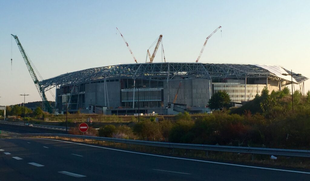 Uma vista de um estádio em construção, com guindastes ao redor da estrutura inacabada, exibindo sua estrutura de aço e base de concreto.