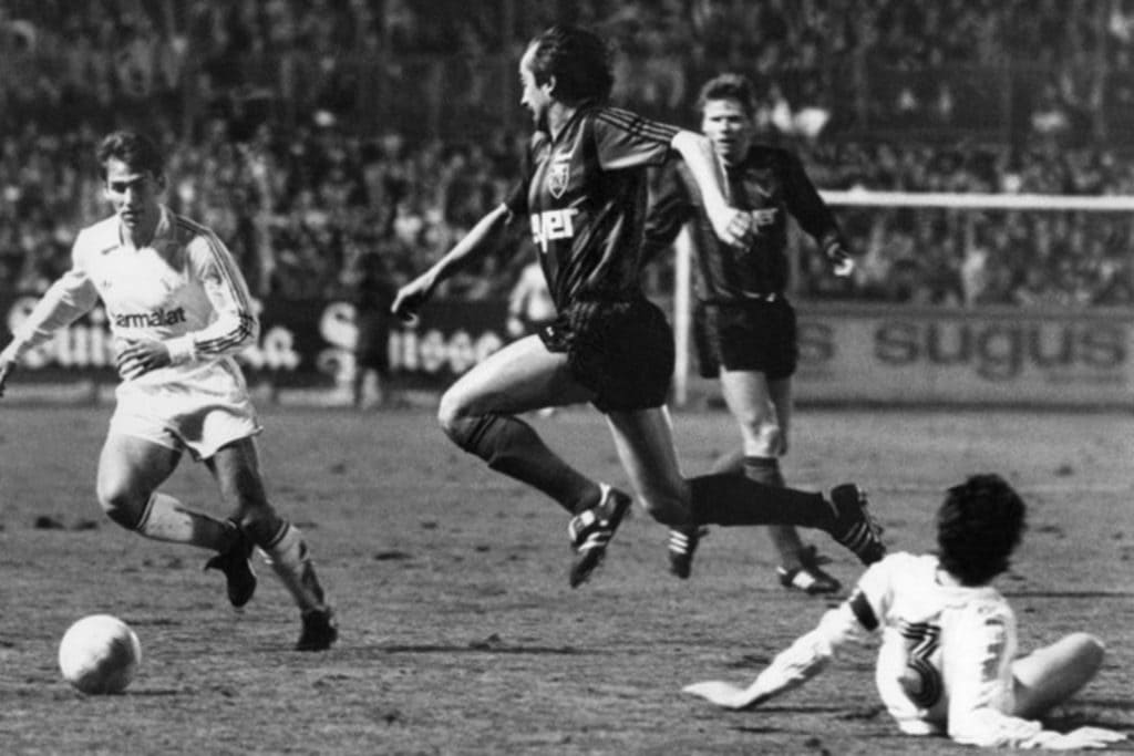 Foto em preto e branco capturando um momento intenso durante uma partida de futebol. Um jogador com uniforme branco está driblando a bola, enquanto os adversários com uniformes escuros o perseguem e defendem.