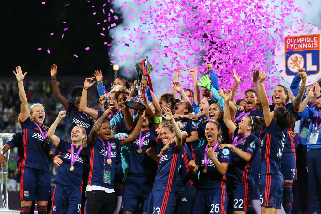 A equipe feminina do Olympique Lyonnais comemora sua vitória, levantando um troféu em meio a confetes rosa e expressões de alegria.