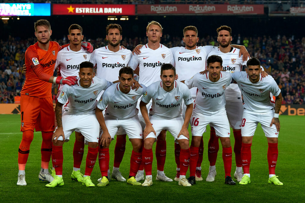 Foto de equipe dos jogadores do Sevilla FC posando no campo antes de uma partida, vestindo seus uniformes branco e vermelho. 