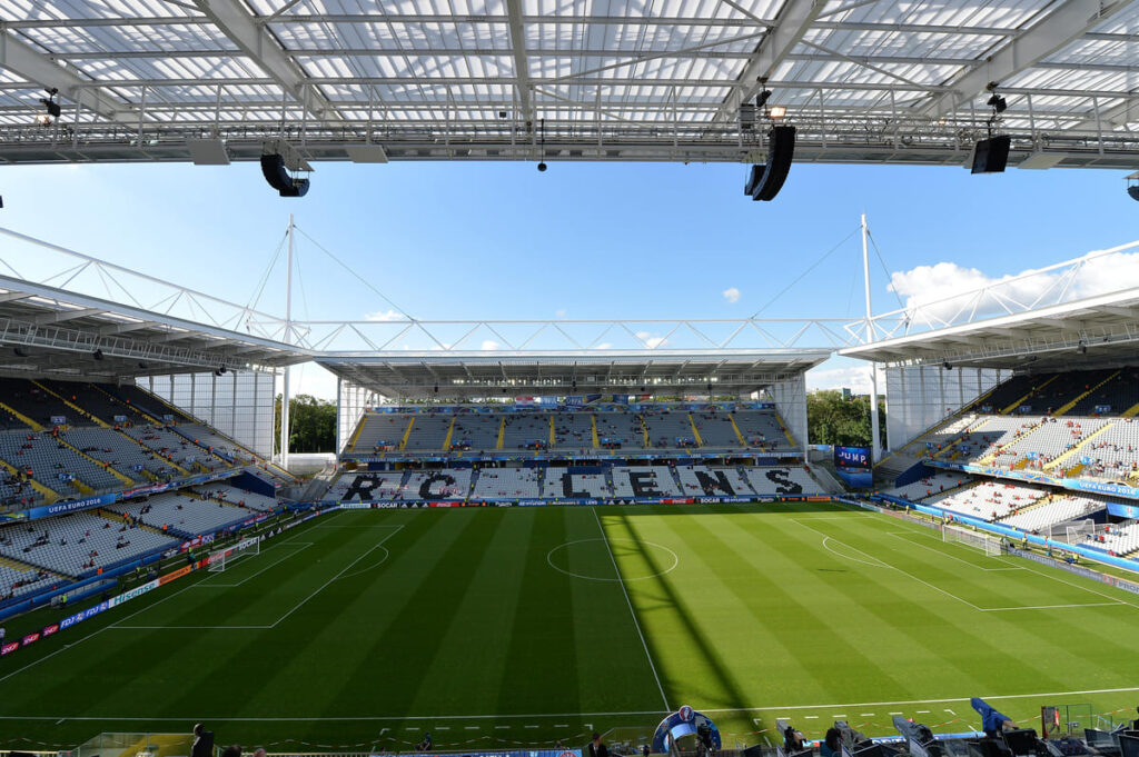 Wide-angle view of a modern football stadium with empty seats and a green pitch, taken during daytime. The stands display the name 'RC LENS'.