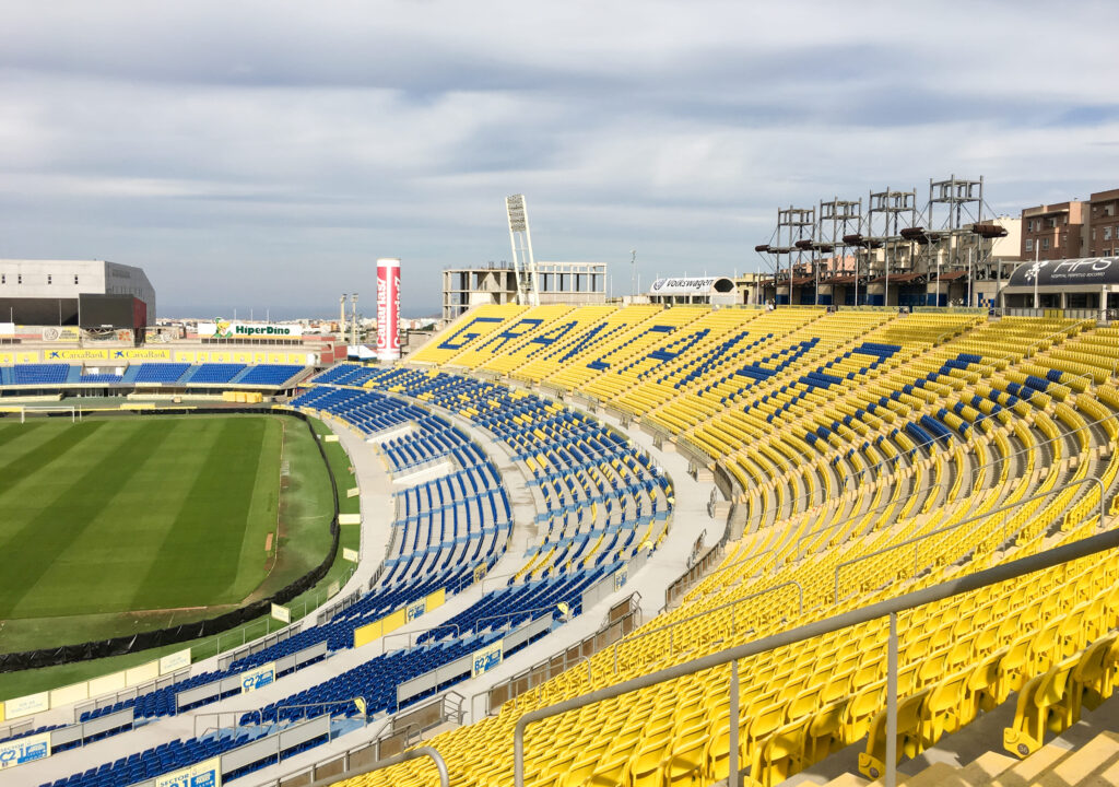 Estadio de Gran Canaria, home of UD Las Palmas, featuring vibrant yellow and blue seating with a clear sky backdrop.