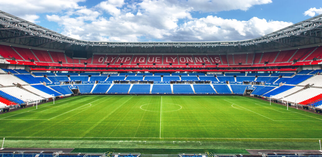 Um estádio de futebol vazio com assentos vermelhos, brancos e azuis, exibindo o letreiro 'Olympique Lyonnais' acima do campo sob um céu parcialmente nublado.