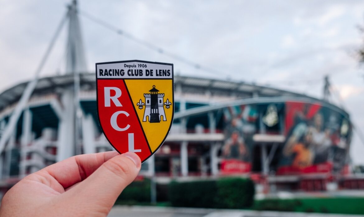 The emblem of the RC Lens football club against the background of a stadium.
