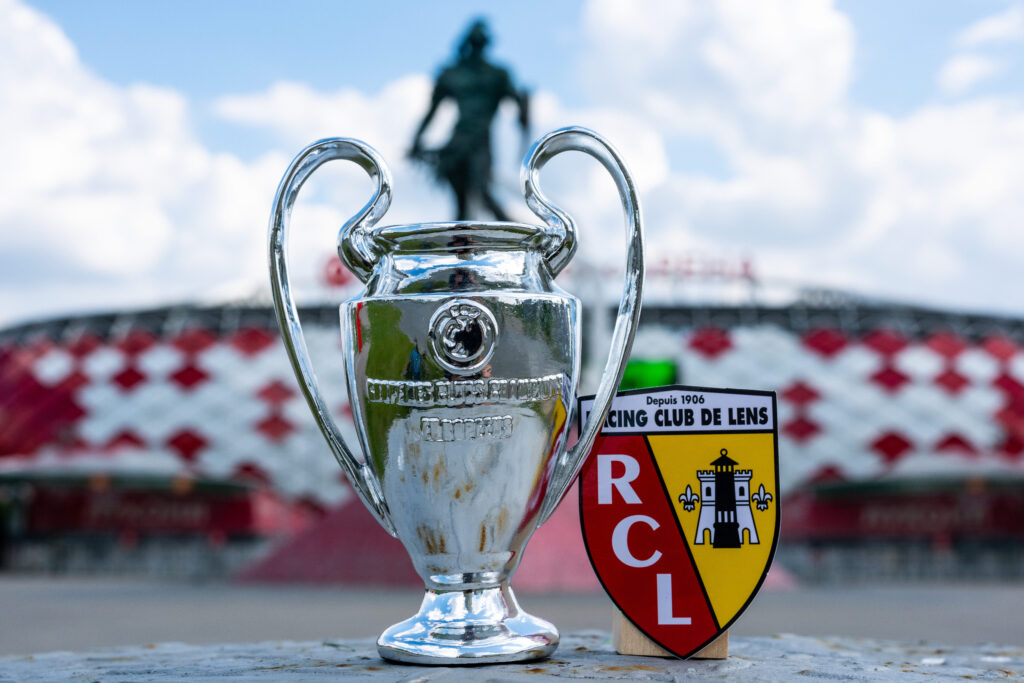 Close-up of a silver Champions League-style trophy next to the Racing Club de Lens (RCL) crest. In the background, the red and white facade of Stade Bollaert-Delelis and a statue of a figure are visible under a bright blue sky with scattered clouds