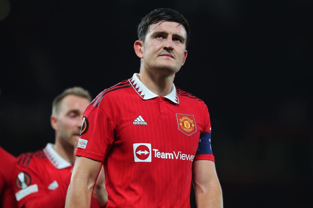 Zdjęcie Harry'ego Maguire'a w koszulce Manchesteru United z opaską kapitana. Skoncentrowany wyraz twarzy zawodnika odzwierciedla determinację.