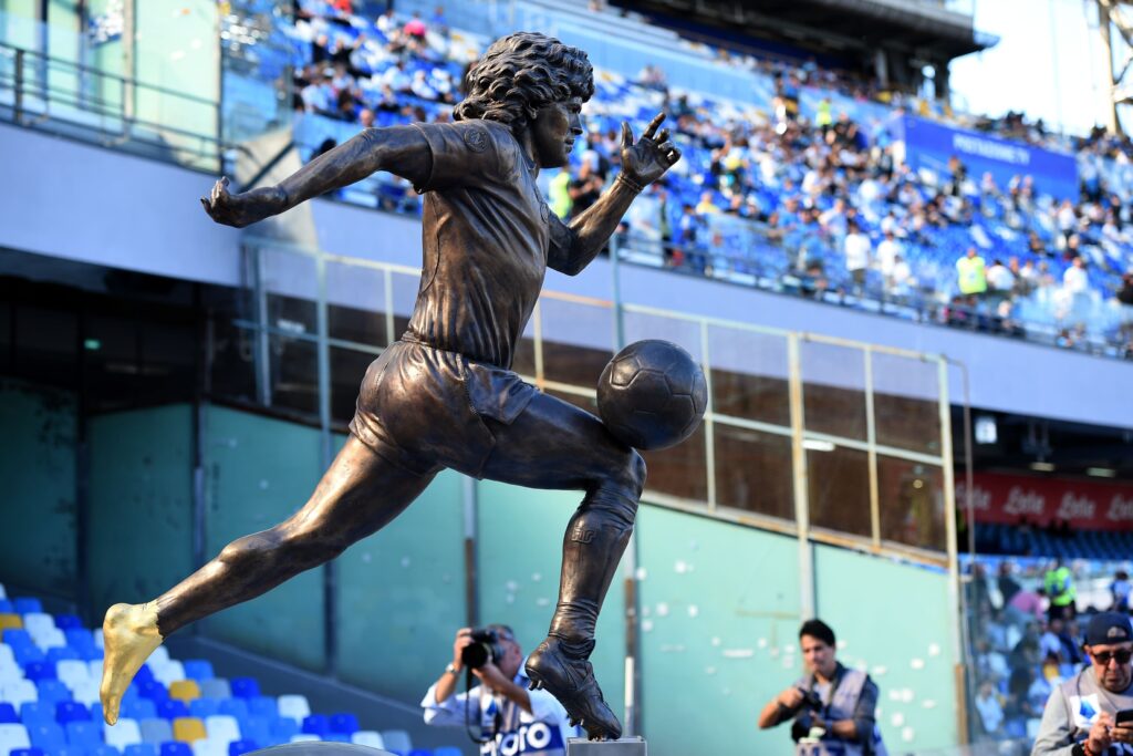 Estátua de bronze de Diego Maradona em uma pose icônica correndo com uma bola de futebol, localizada em um estádio com assentos azuis e brancos.