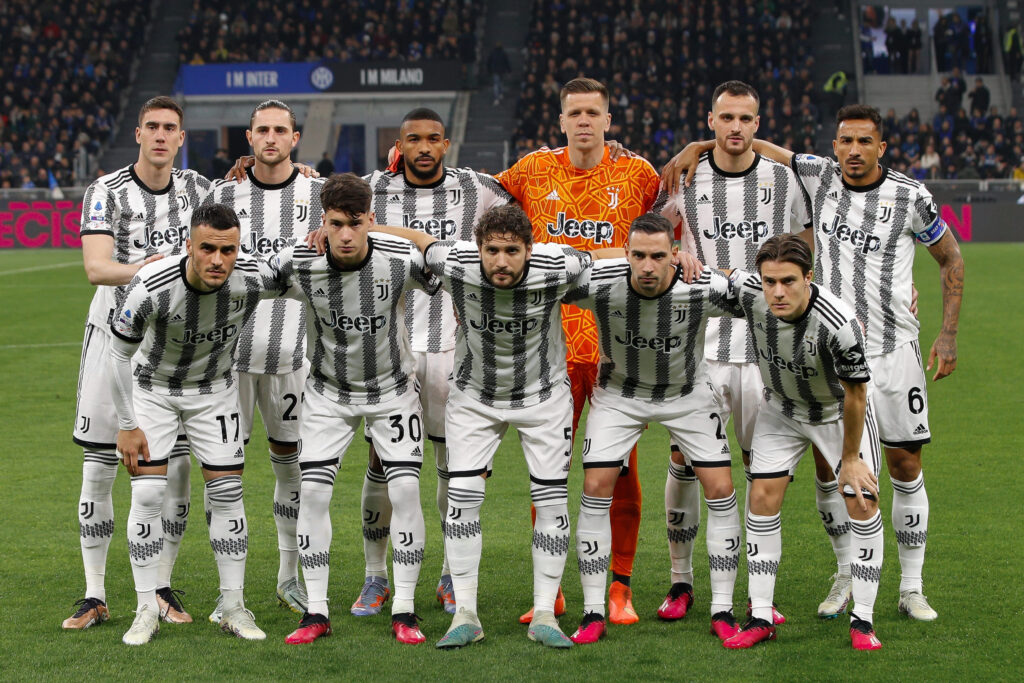 Foto de equipe dos jogadores da Juventus posando no campo antes de uma partida, vestindo seus uniformes clássicos listrados em preto e branco. 