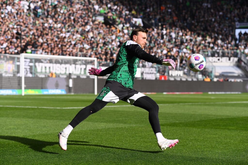 Um goleiro em ação durante uma partida, vestido com um uniforme verde e preto e luvas rosas, focado em chutar a bola. 