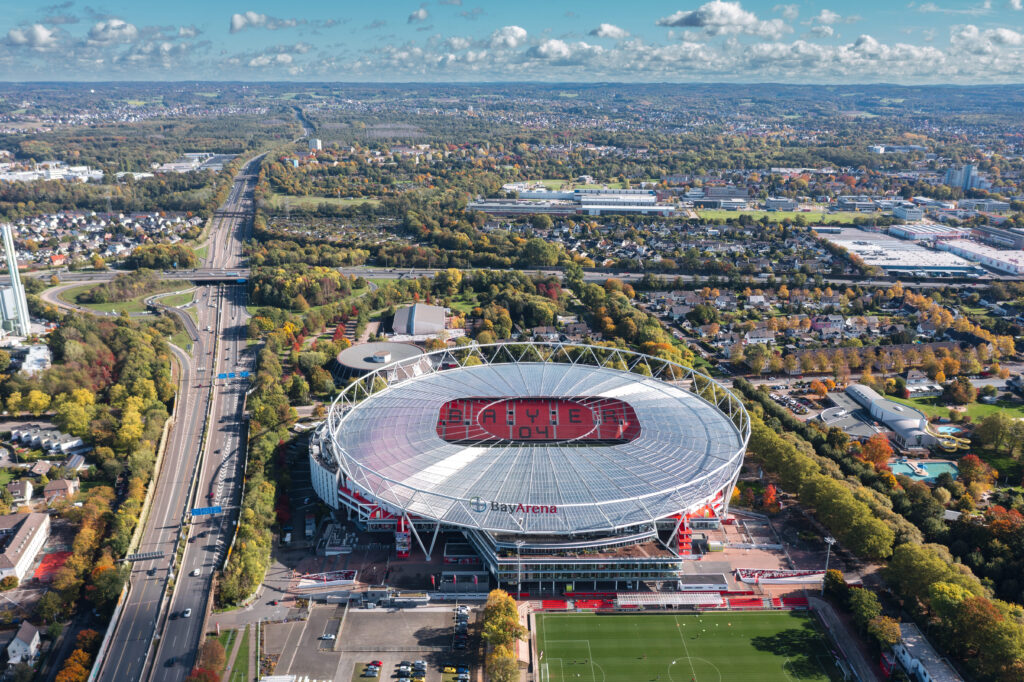 An aerial view of BayArena, the home stadium of Bayer Leverkusen, surrounded by lush greenery and urban landscapes in Leverkusen, Germany.