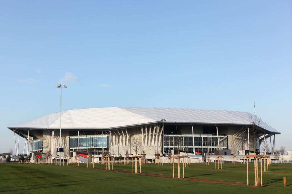 Uma vista externa diurna do Estádio Groupama, mostrando seu design moderno com um telhado branco e uma fachada de vidro.