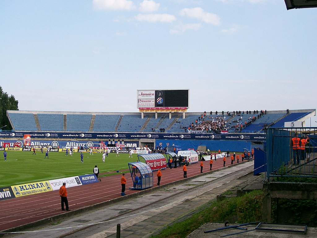 View of Stadion Maksimir in Zagreb, Croatia, showing a football match in progress, a sparsely populated south stand, a digital scoreboard, and security personnel along the running track.