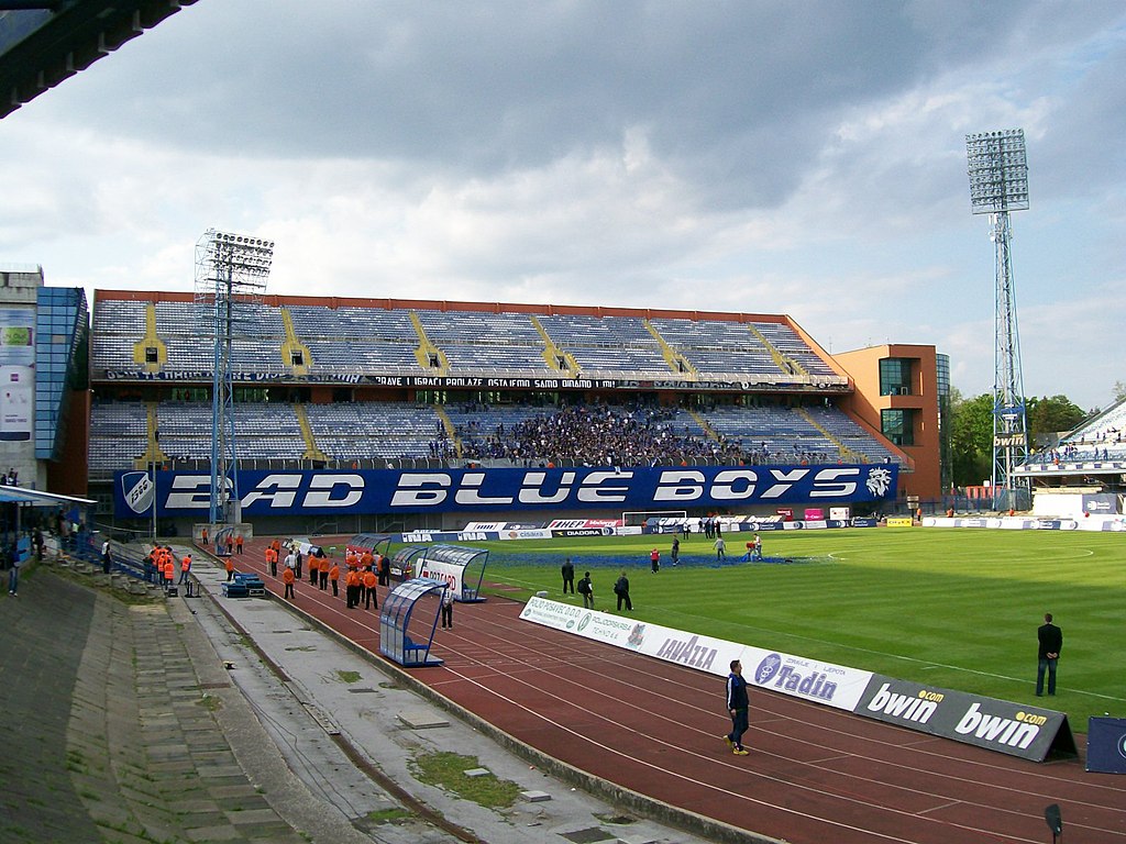 View of Stadion Maksimir in Zagreb, Croatia, featuring the "Bad Blue Boys" supporters' section, a running track around the pitch, and floodlights under a cloudy sky.