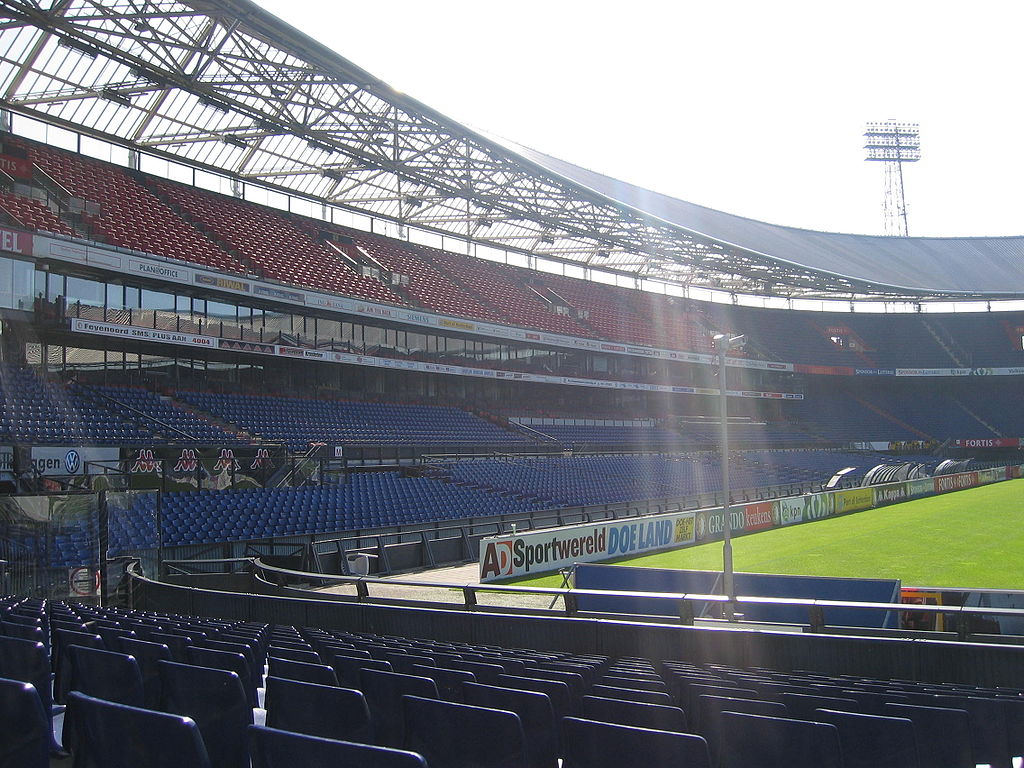 A view of De Kuip Stadium in Rotterdam, featuring red and blue seating, a partially covered roof, and a sunlit football pitch.