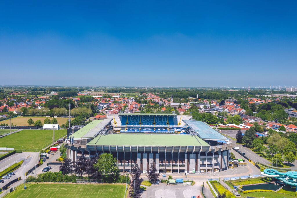 A high-angle shot of Jan Breydel Stadium, with its blue and black seats, and the field visible below, with a clear blue sky above.