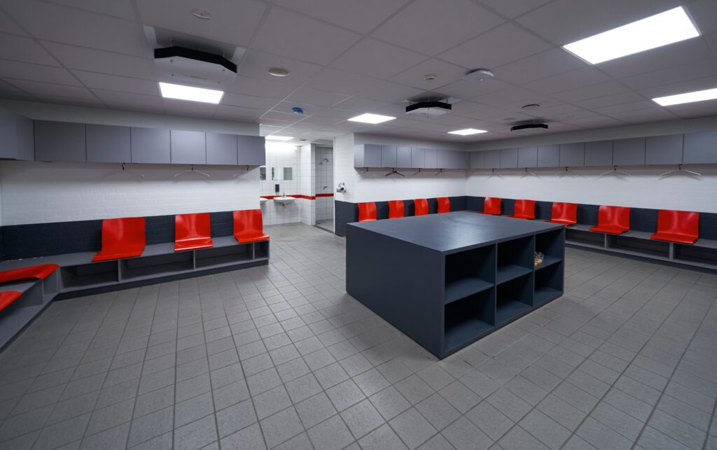 Modern football locker room with red and grey seating, storage compartments, and bright overhead lighting.