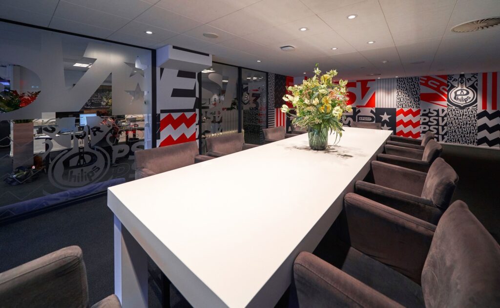 Modern meeting room with a long white table, plush chairs, and a floral centerpiece, featuring PSV Eindhoven-themed wall decor and frosted glass partitions.