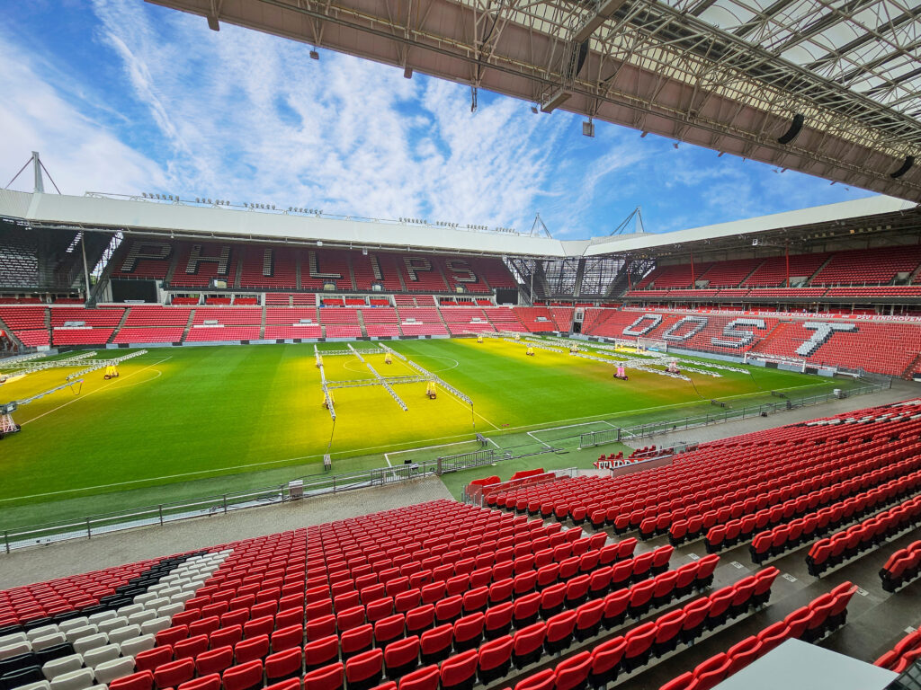 Philips Stadion, home of PSV Eindhoven, featuring red seating, a well-maintained pitch with artificial grow lights, and a clear blue sky.