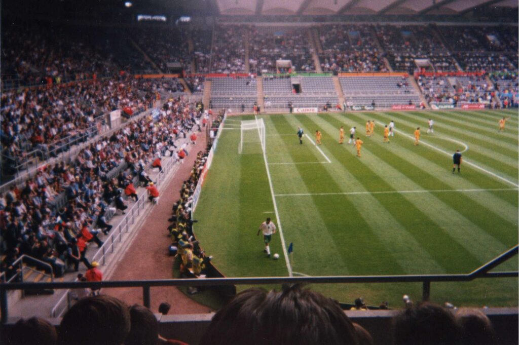 Football match at a stadium during Euro 96, with a player preparing to take a corner kick, teams in action on a striped green pitch, and a crowd of spectators in the stands.