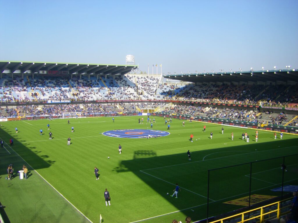 A shot of a soccer game at Jan Breydel Stadium, featuring the Club Brugge players in their blue and black uniforms, standing around the referee as he reviews a VAR decision.