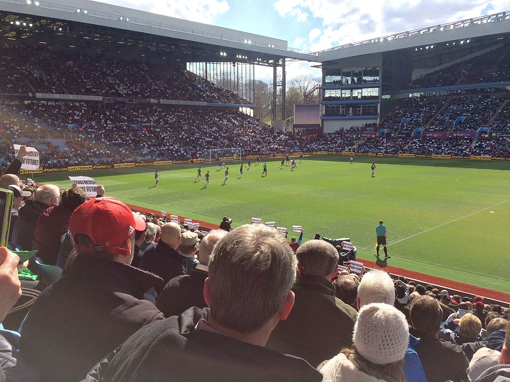 View from the stands at Villa Park stadium during a football match, with a crowd of spectators, some holding protest signs, and players in action on the pitch.