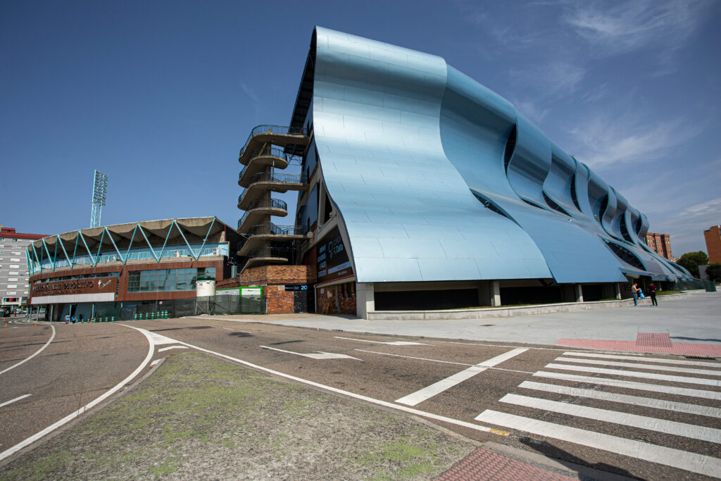 A modern stadium with a striking blue, wave-like façade and an adjacent traditional structure, set against a clear sky.