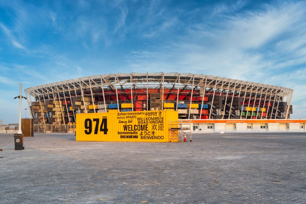 A frontal view of Stadium 974 in Doha, Qatar, constructed from shipping containers, under a bright blue sky with scattered clouds.