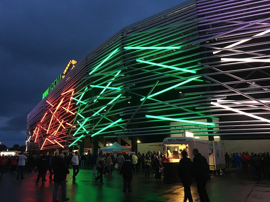 Night view of WWK Arena in Augsburg, Germany, featuring an illuminated facade with red, green, and white lights, as a crowd gathers outside near food stands.