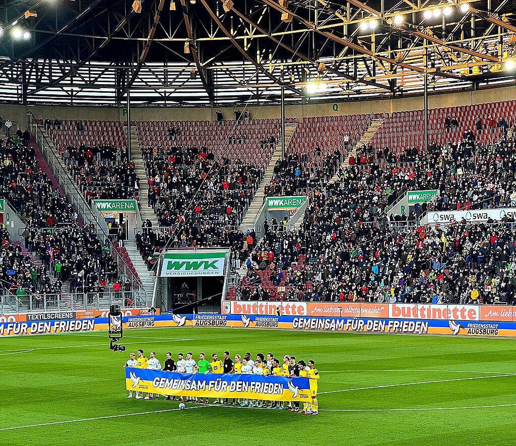 Players from FC Augsburg and Borussia Dortmund stand together holding a "Gemeinsam für den Frieden" ("Together for Peace") banner before a Bundesliga match at WWK Arena, with fans filling the stands.