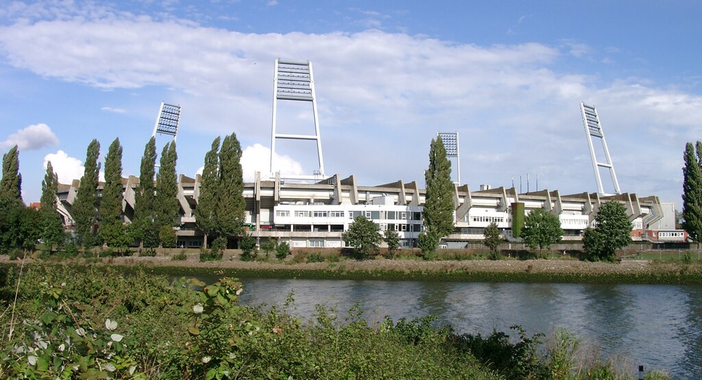 Exterior view of Weserstadion in Bremen, Germany, with its distinctive floodlight towers, surrounded by trees and the Weser River under a partly cloudy sky.