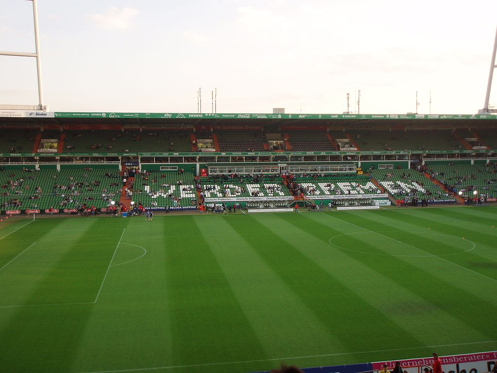 Interior view of Weserstadion in Bremen, Germany, showing green seating, a well-maintained pitch, and fans arriving before a match, with "WERDER BREMEN" spelled out in the stands.