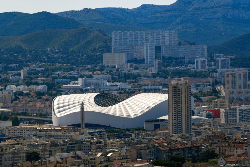 A panoramic view of Marseille, France, featuring the modern, wave-like roof of the Stade Vélodrome surrounded by the city's dense urban landscape and backed by rugged hills.