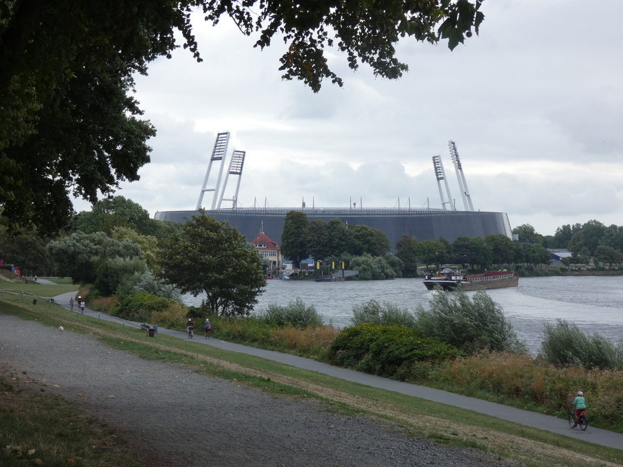 Scenic view of Weserstadion in Bremen, Germany, with its distinctive floodlight towers, surrounded by lush greenery and the Weser River, as people walk and cycle along the riverside path.