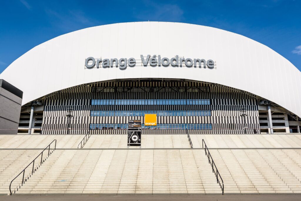 Front view of the Orange Vélodrome stadium, featuring a modern arched design, bold signage, and wide entrance steps under a clear blue sky.