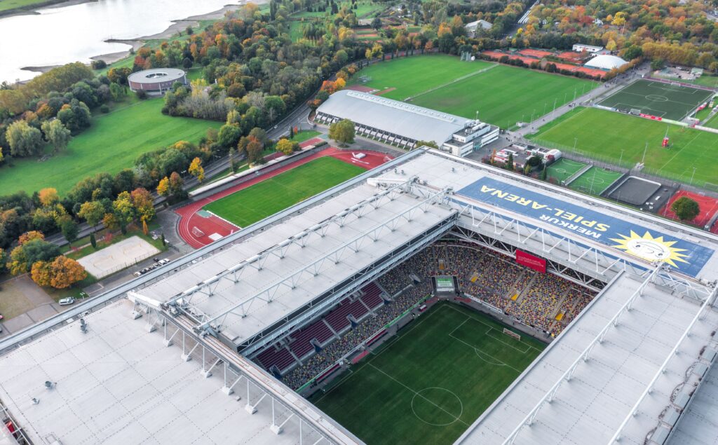 An aerial view of the Merkur Spiel-Arena in Düsseldorf, Germany, showing the stadium filled with spectators, surrounded by green fields, training grounds, and a river in the background.