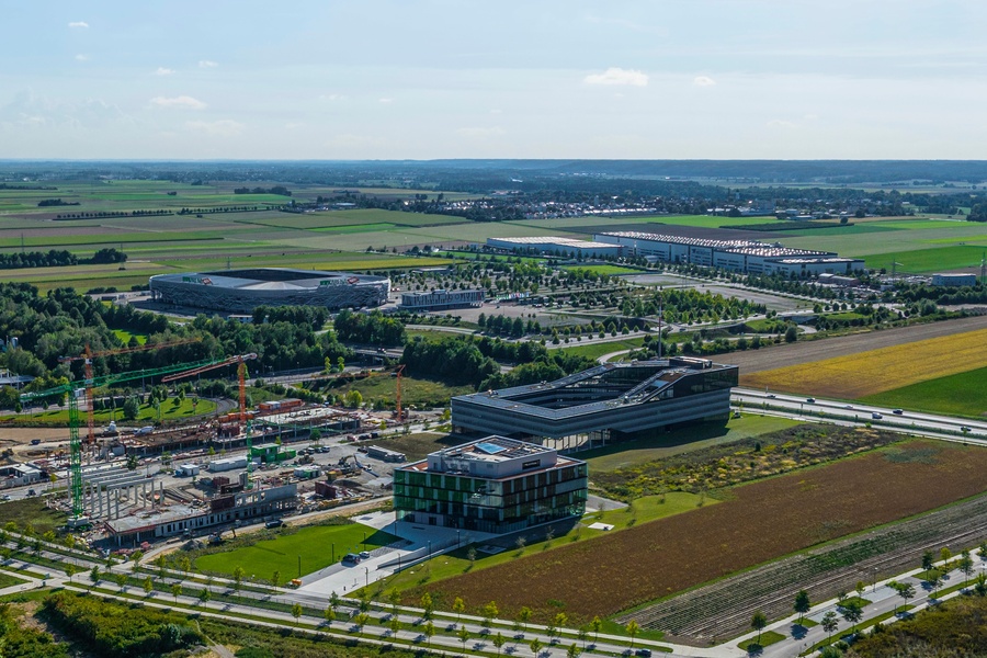 Aerial view of a modern stadium surrounded by open fields, roads, and construction sites, with additional buildings and infrastructure in the background under a clear sky.