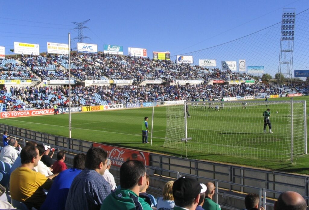 View from the stands at Coliseum Alfonso Pérez stadium in Getafe, showing a football match in progress, with a goalkeeper near the net, spectators in blue seats, and advertisement banners around the pitch.