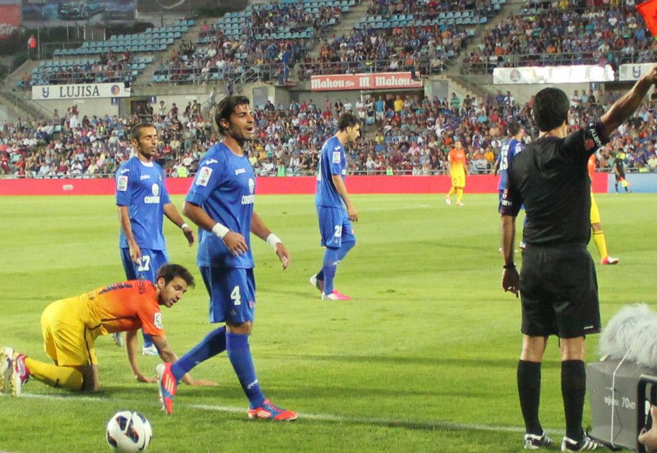 A football match at Coliseum Alfonso Pérez stadium, with a referee raising the flag, a Getafe player reacting, and a Barcelona player on the ground, while the crowd watches from the stands.