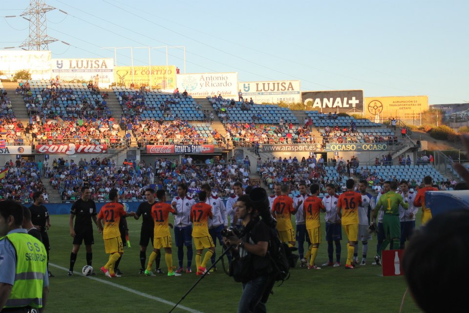 Players from two teams greet each other before a football match at Coliseum Alfonso Pérez stadium in Getafe, with spectators in blue seats and advertisement banners in the background.