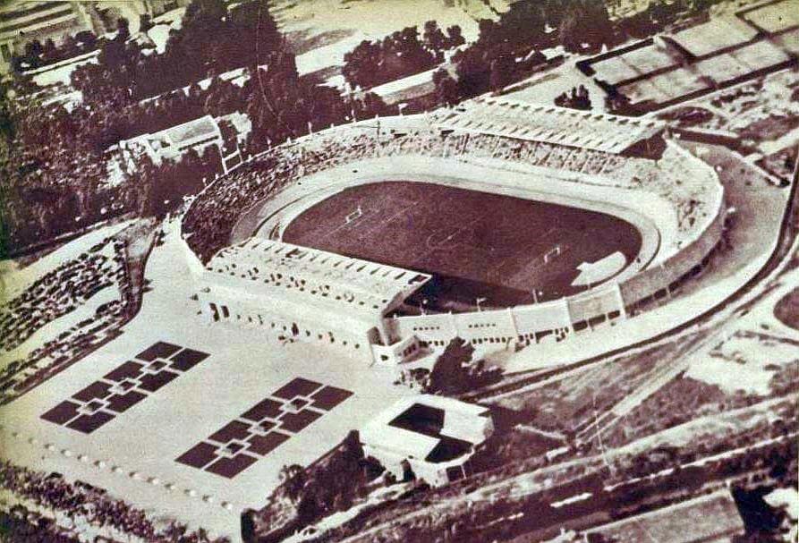 Vintage aerial view of an oval stadium with grandstands, a crowded audience, and surrounding open spaces.